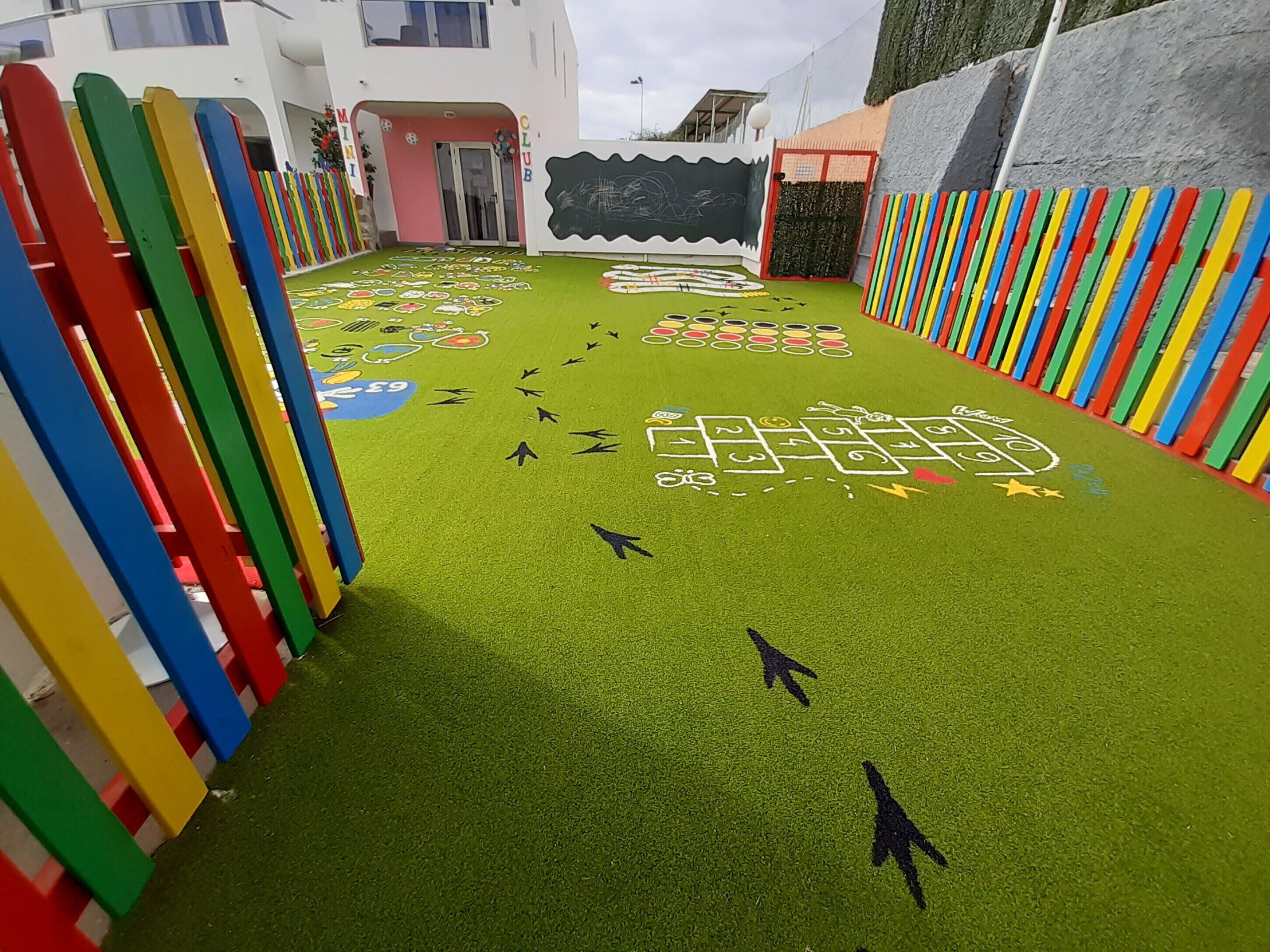 Zona infantil con un juego de rayuela, vallas coloridas y la entrada de un edificio.