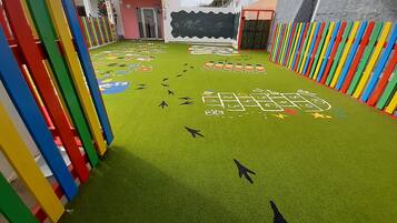 Zona infantil con un juego de rayuela, vallas coloridas y la entrada de un edificio.