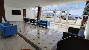 A spacious lobby with a tiled floor, blue and black seating, a flat-screen TV, and large windows offering a view of a residential area. - Apartments Turbo Club (San Bartolomé de Tirajana)