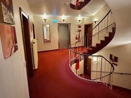 Un pasillo de hotel con alfombra roja, una escalera en espiral y una puerta al final.