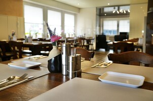 Un área de comedor con mesas dispuestas para dos personas, que incluye un jarrón con flores, un salero y pimentero, y un plato blanco.