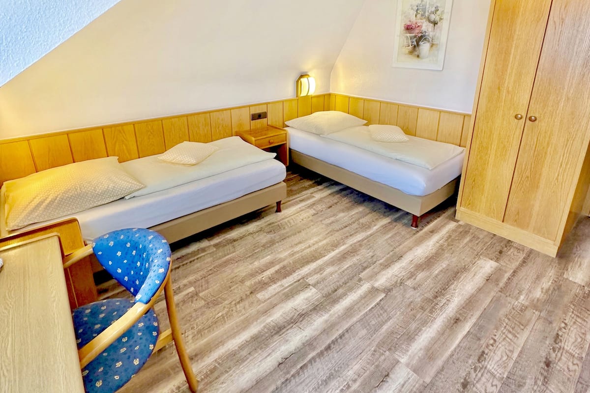 A small, cozy room with two single beds, a wooden wardrobe, a desk with a chair, and a wall-mounted light.