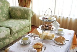 Un soporte de tres niveles con diversos pasteles y tartas, una tetera blanca y tazas de té sobre una mesa en una habitación con un sofá de estampado verde.