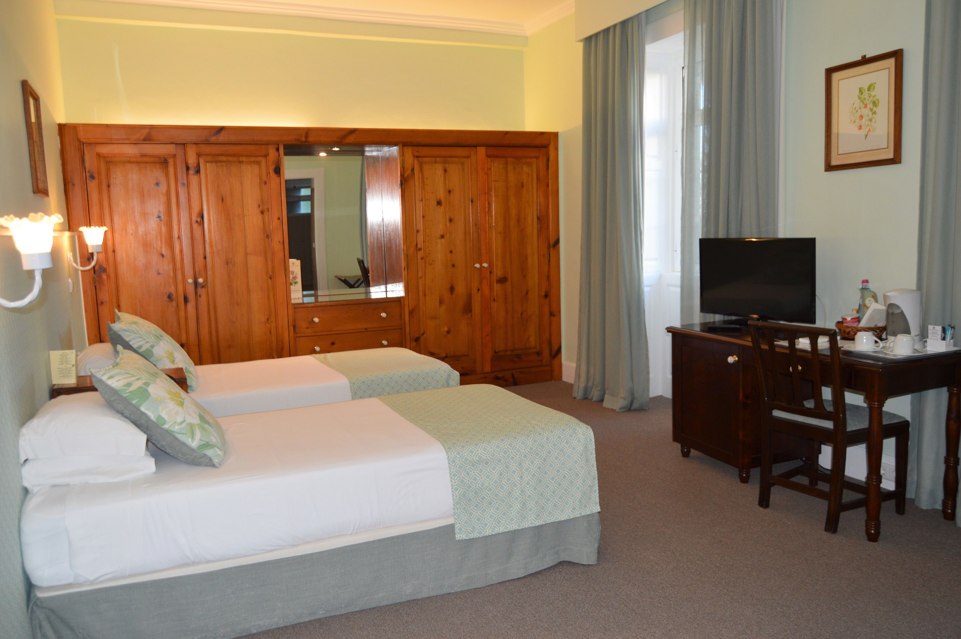 A hotel room with two single beds, a wooden wardrobe, a desk with a television, and a chair.
