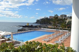 A pool area with lounge chairs and umbrellas, overlooking the sea and a coastal cityscape.