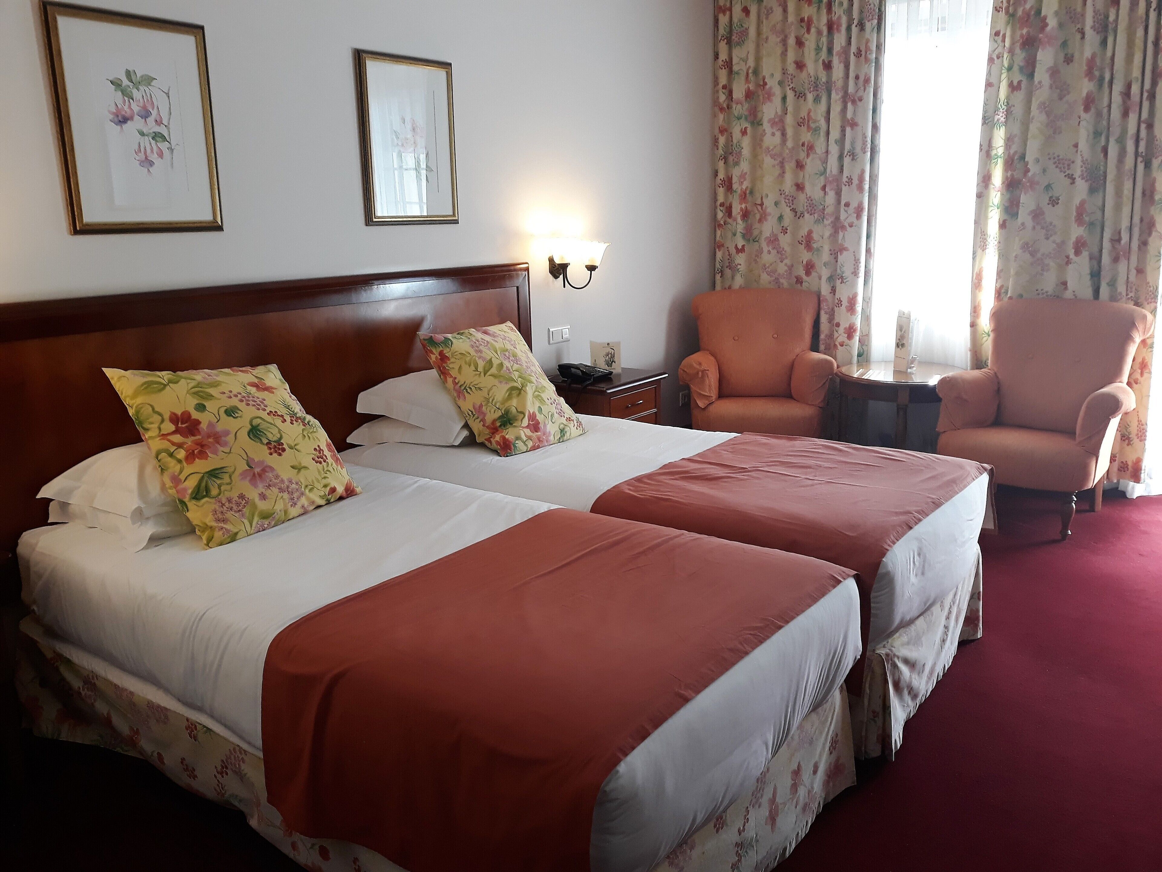 Ein Hotelzimmer mit zwei Betten, Kissen mit Blumenmuster, einem Holzkopfende, einem Nachttisch, einer Lampe, einem Sessel und einem Fenster mit floralen Vorhängen.