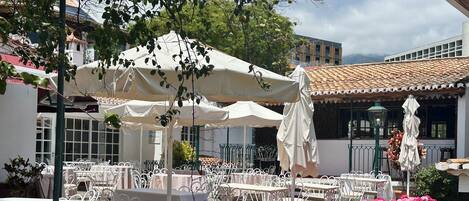 An outdoor café with white tables and chairs, surrounded by a stone wall and vibrant flowers.
