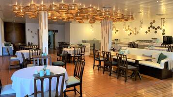 A spacious dining area with multiple tables and chairs, a central seating area with a sofa, and a kitchen in the background.