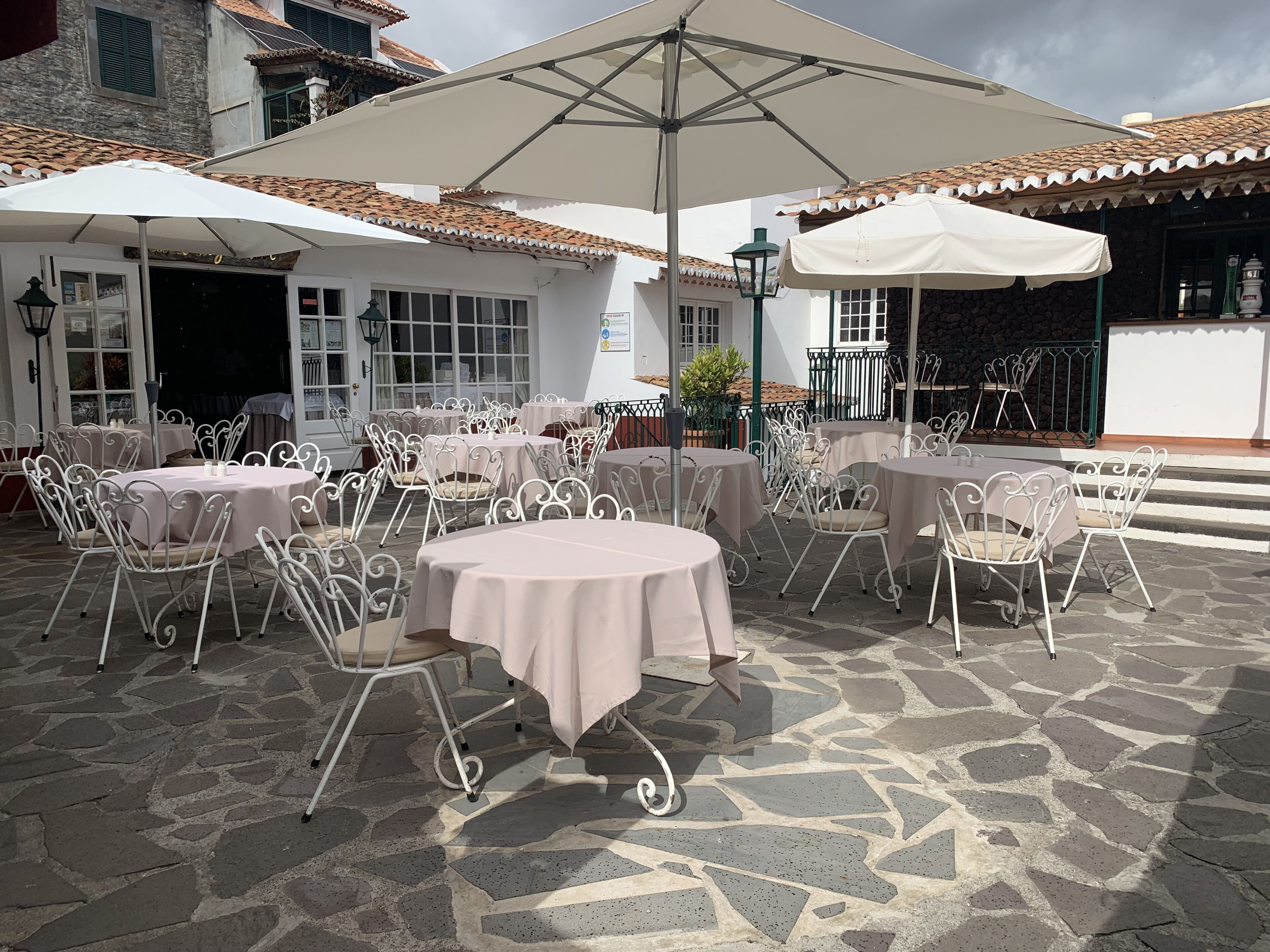 Un patio al aire libre con mesas redondas y sillas de metal blanco, cubierto por grandes sombrillas.