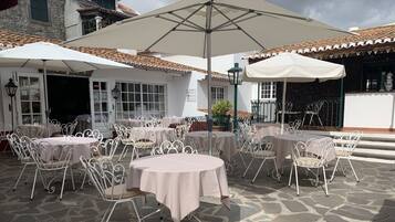 Un patio al aire libre con mesas redondas y sillas de metal blanco, cubierto por grandes sombrillas.