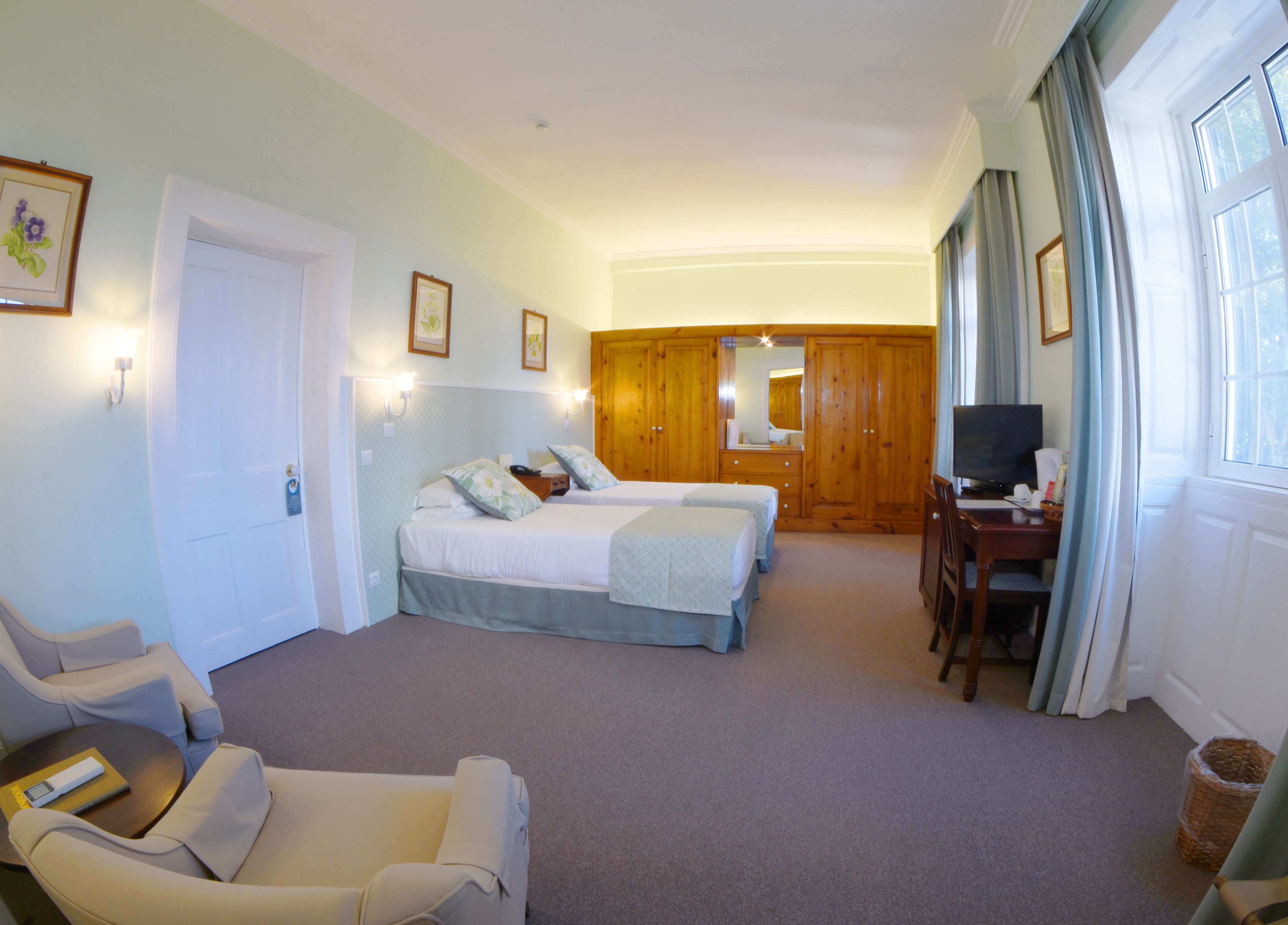 A hotel room with two beds, a desk with a television, a chair, and a large window with curtains.