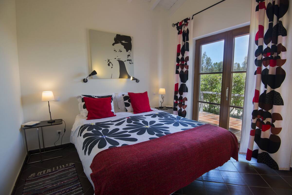 A bedroom with a bed, two bedside tables, a wall-mounted lamp, and a window with curtains.