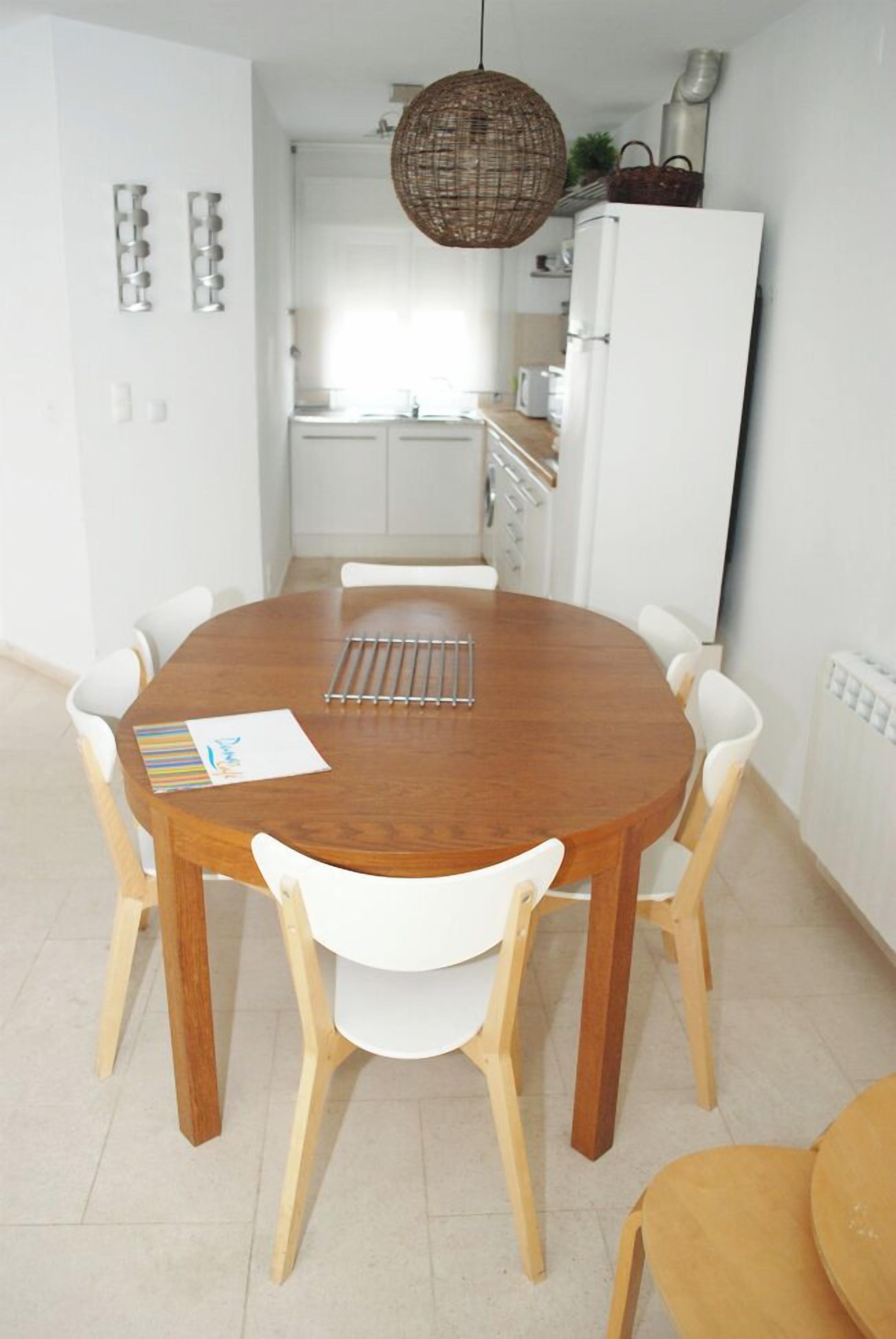 A dining area with a wooden table and white chairs, a hanging light fixture, and a kitchen in the background.