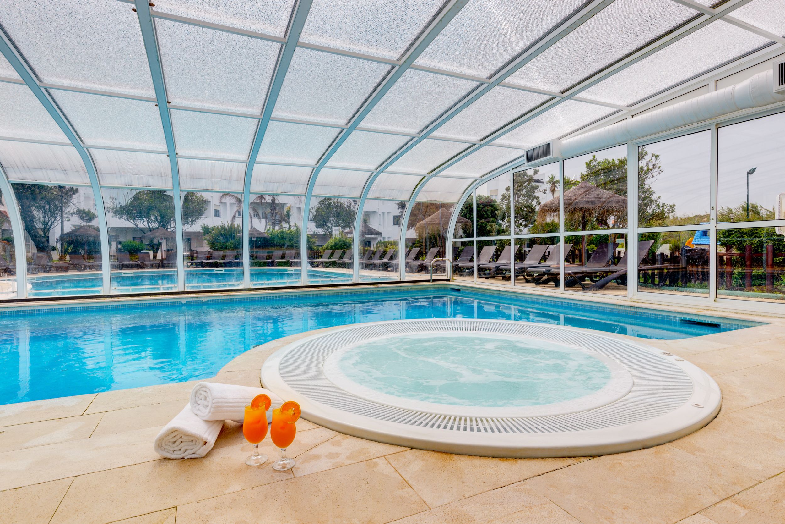 En indendørs swimmingpool med jacuzzi, liggestole og et glastag.
