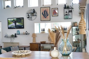 A cozy living room with a television, framed artwork, shelves with bottles, and a wicker lamp.