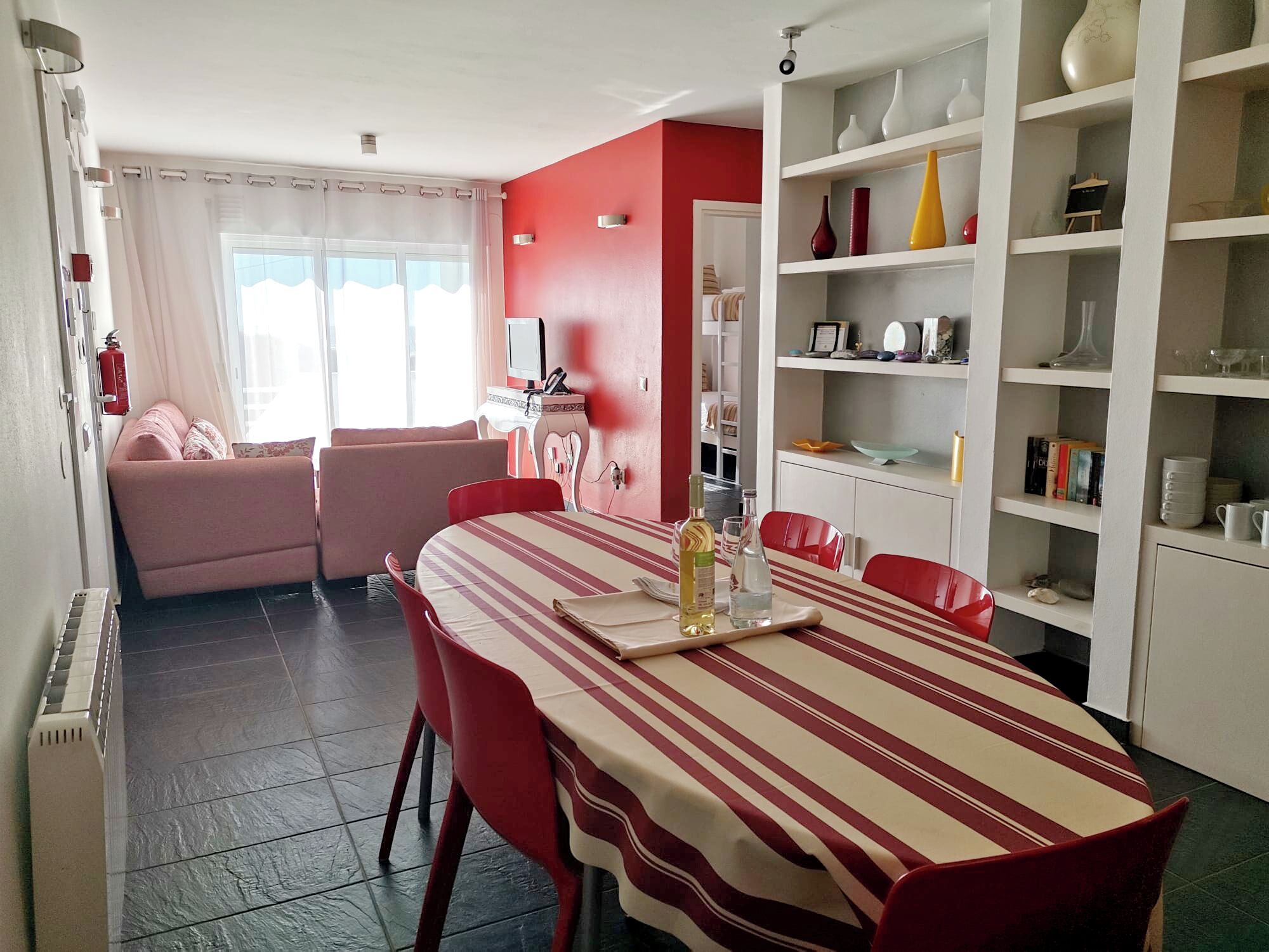 Un coin repas avec une nappe à rayures, des chaises rouges, un canapé et une bibliothèque contenant divers objets.