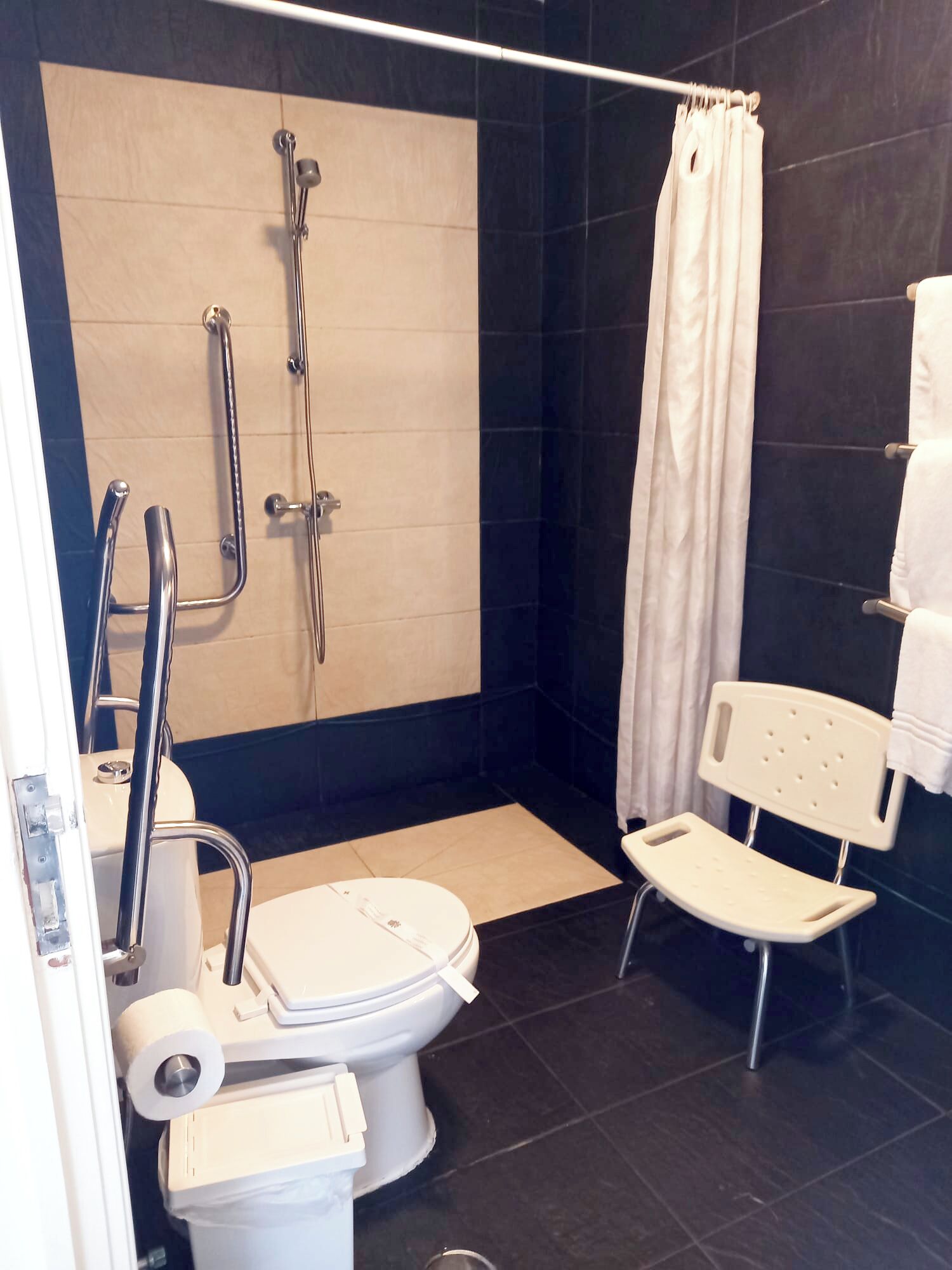 A modern bathroom with a walk-in shower, a toilet with a bidet attachment, a white chair, and a towel rack.