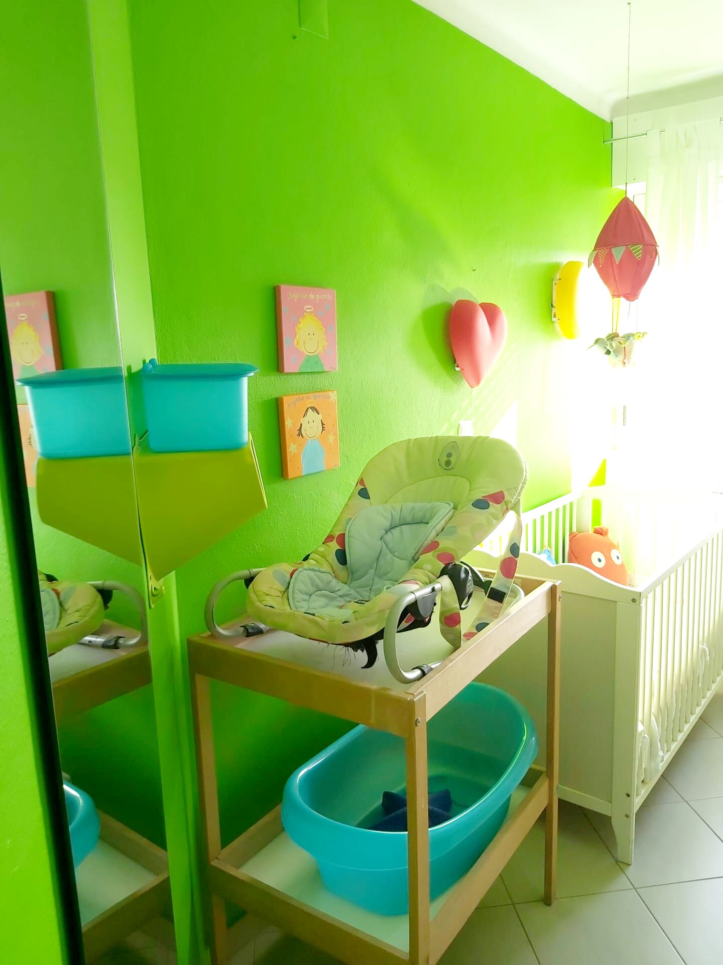 A room with a green wall, a baby changing station, a crib, and a window with curtains.