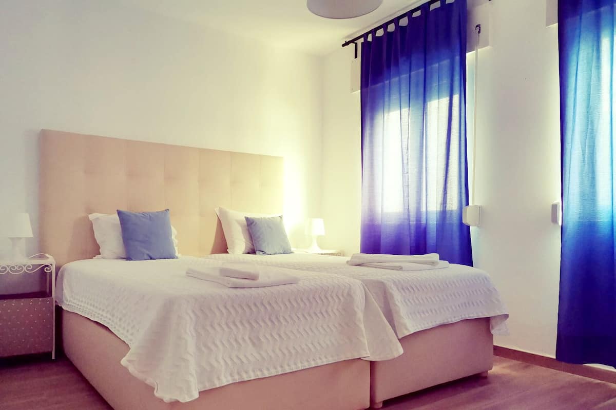 A double bed with a wooden headboard, two pillows, a folded towel, and a bedside table with a lamp.