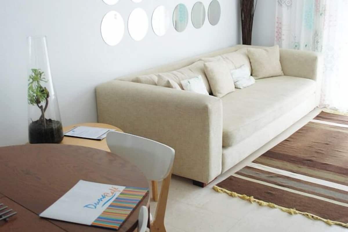 A modern living room with a beige sofa, a round dining table with chairs, and a decorative mirror wall.