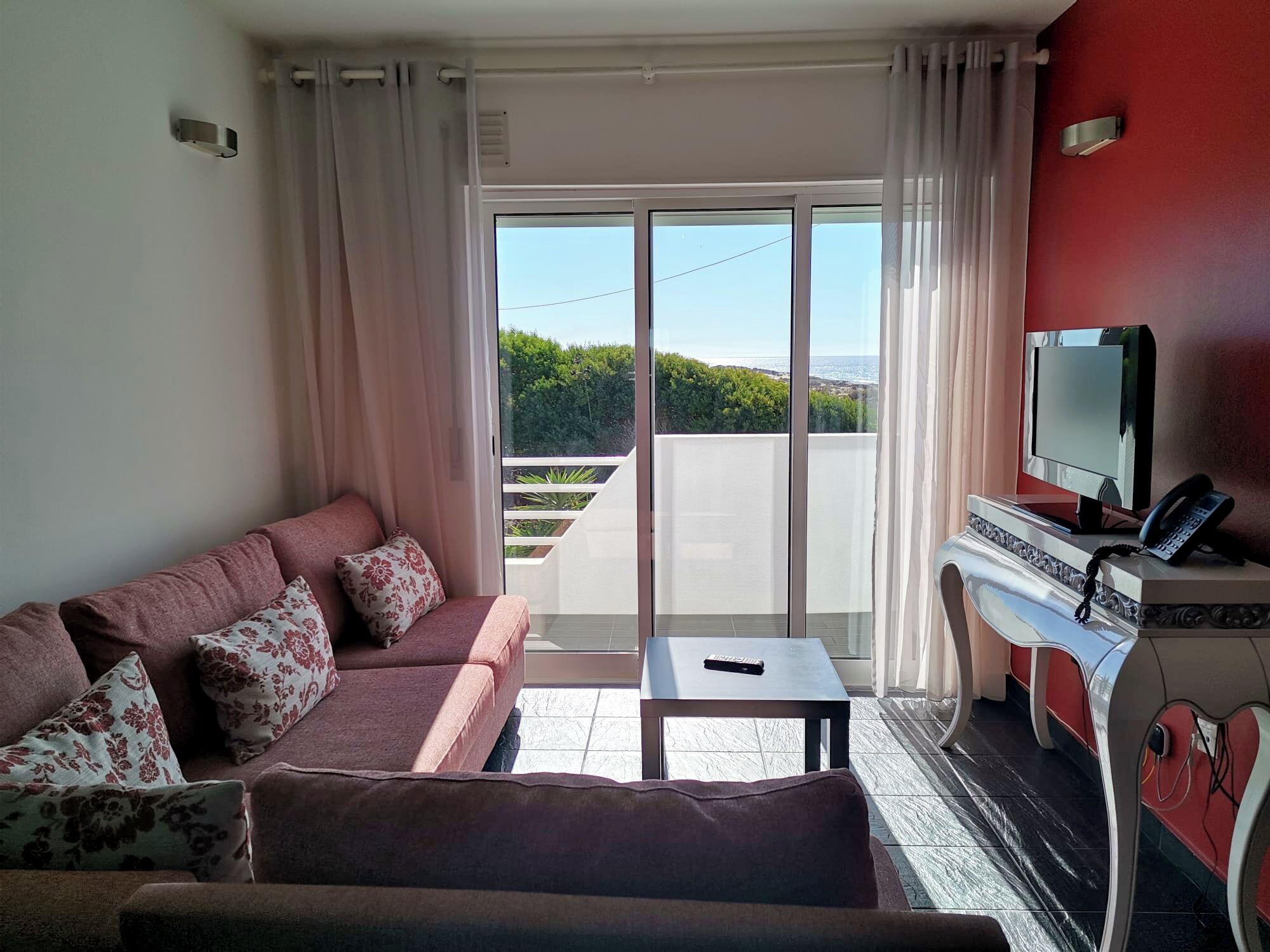 A living room with a sofa, a coffee table, a TV, and a balcony with a view of greenery and the sea.