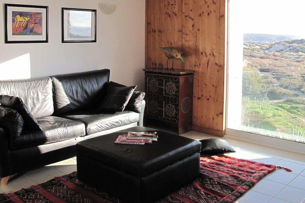 A living room with a leather sofa, a coffee table with magazines, a patterned rug, and a view of a landscape through a large window.