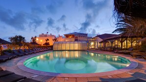 A swimming pool with lounge chairs, a glass-enclosed structure, and a building with a red roof in the background. - Duna Parque Beach Club - Duna Parque Resorts & Hotels (Odemira)