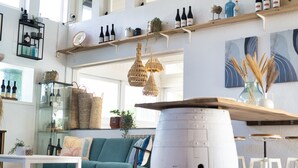 A modern living room with a teal sofa, white coffee table, and a large white barrel. There are shelves with wine bottles and decorative items, and a hanging wicker light fixture. - Duna Parque Beach Club - Duna Parque Resorts & Hotels (Odemira)