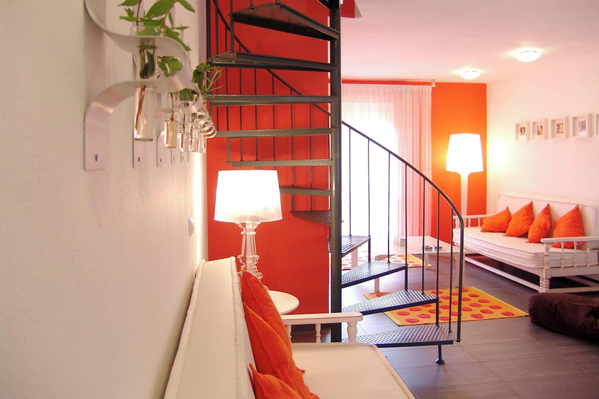 A modern living room with a white sofa, orange cushions, a spiral staircase, and a plant on the wall.