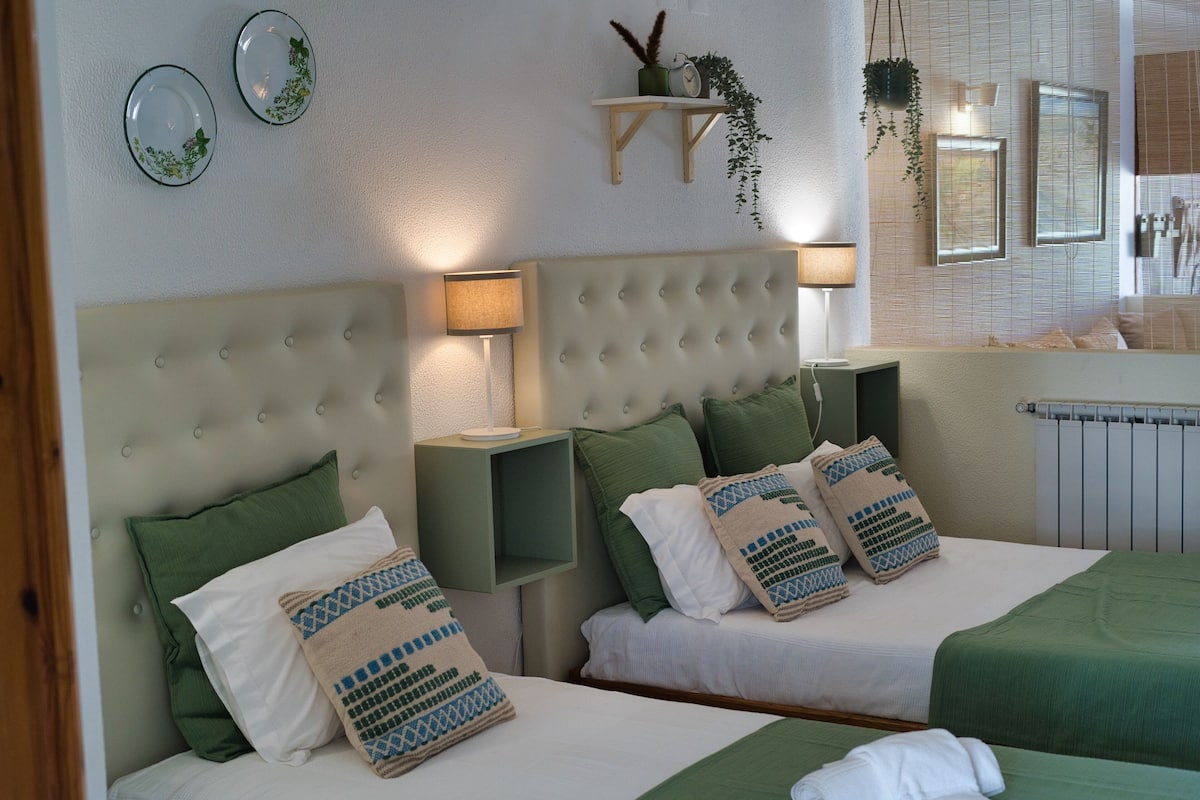 A hotel room with two beds, green and white bedding, a tufted headboard, bedside lamps, a small shelf with plants, and a mirror above the bed.