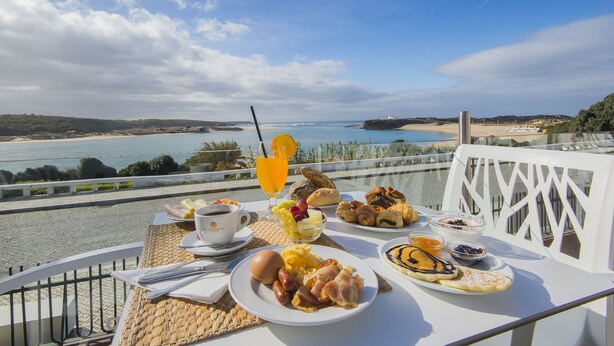 A table set with breakfast items including croissants, eggs, and fruit, overlooking a coastal view. - Duna Parque Beach Club - Duna Parque Resorts & Hotels (Odemira)