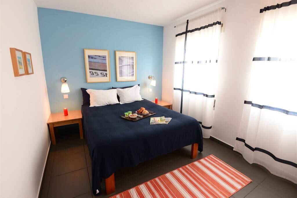 Hotellihuone, jossa on sänky, kaksi kehystettyä kuvaa seinällä, yöpöytä lampun kanssa ja ikkuna, jossa on verhot.