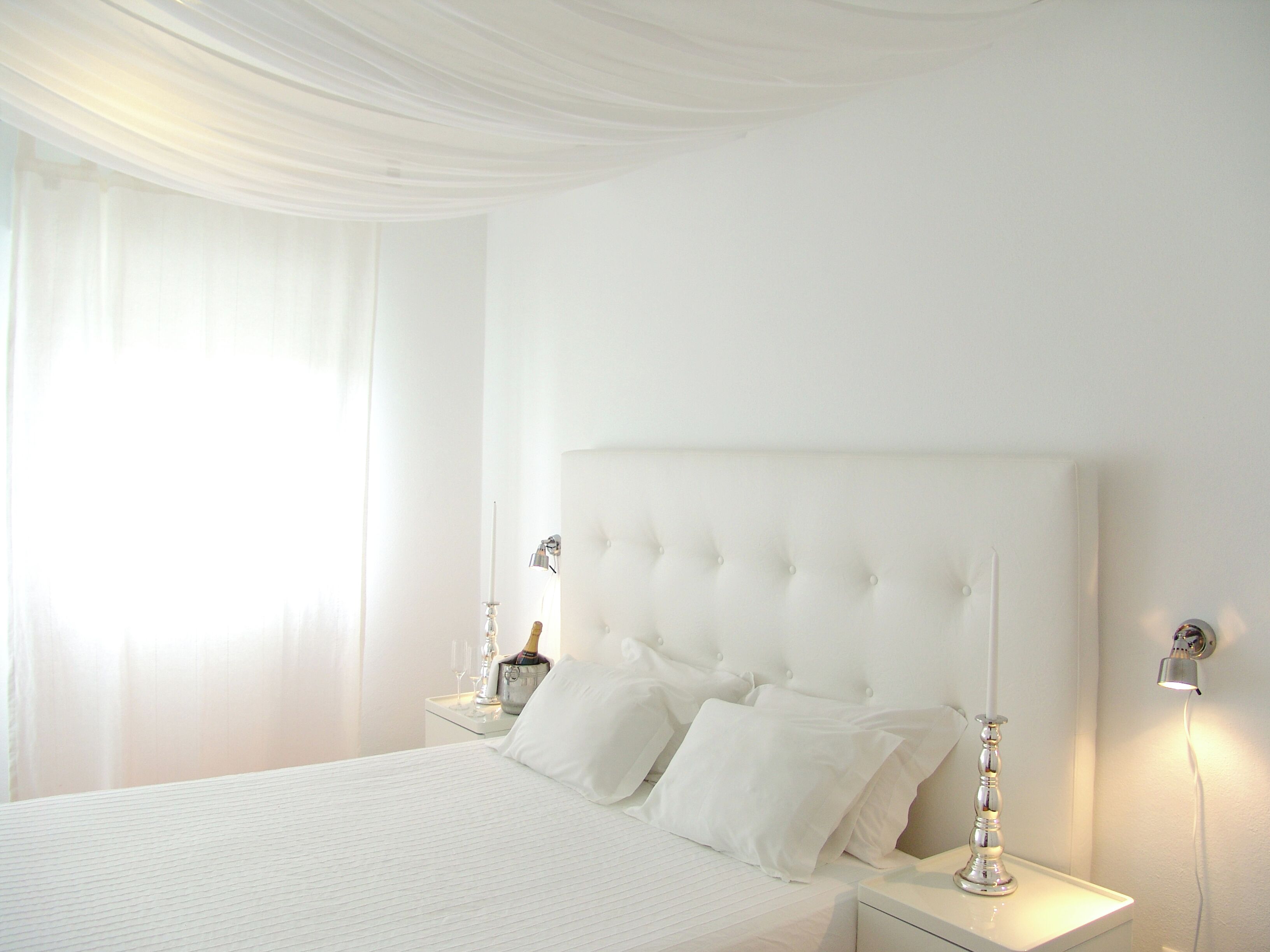 A white bedroom with a tufted headboard, a bedside table with a lamp, and a nightstand with a bottle and glasses.
