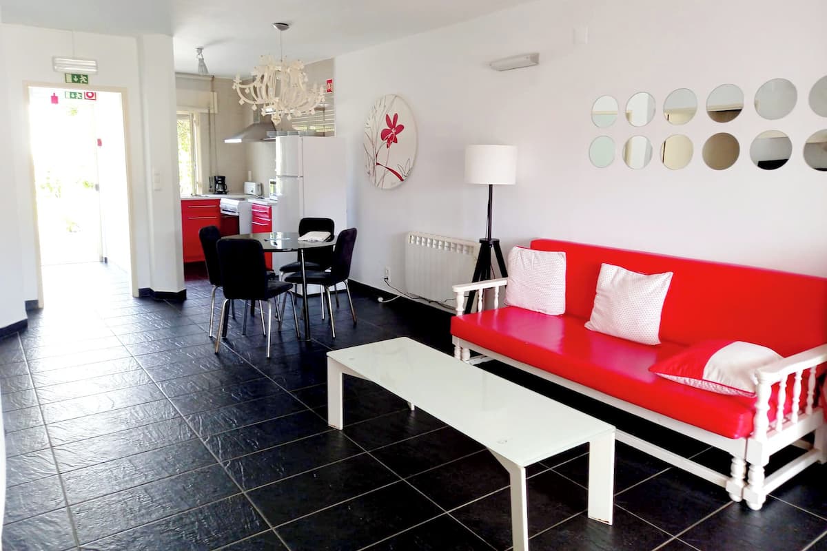 A modern living room with a red sofa, white bench, and black chairs. A dining area with a table and chairs is visible in the background.