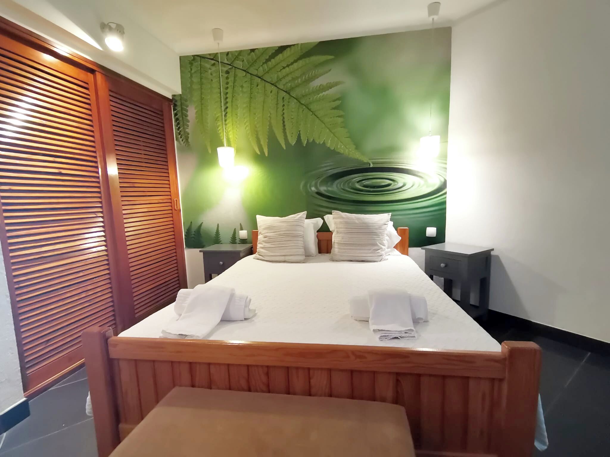 Una cama bien hecha con sábanas y almohadas blancas, un cabecero de madera y una pared con un mural de una hoja verde grande.