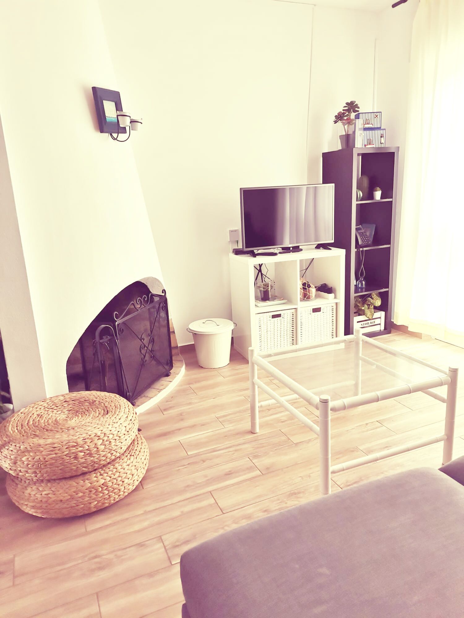 A living room with a fireplace, a television on a stand, a bookshelf, and a wicker pouf.