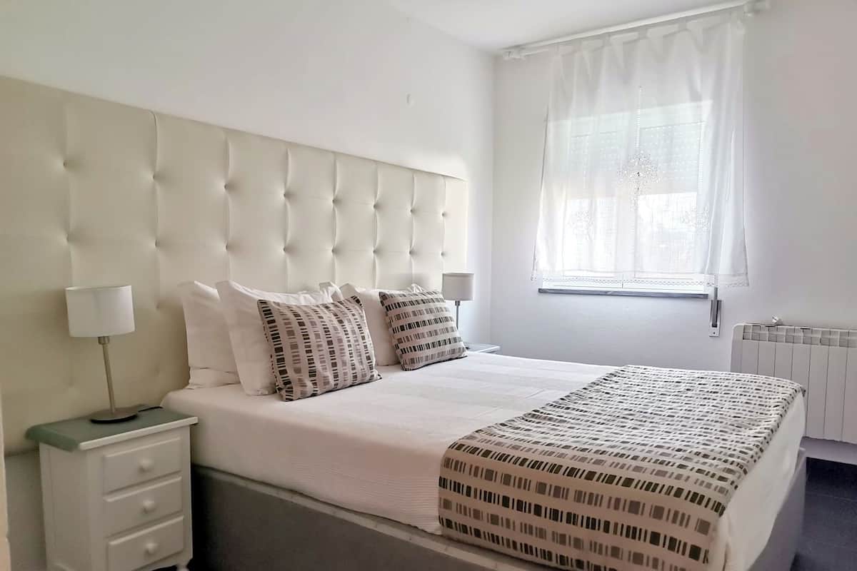 A bedroom with a tufted headboard, a bed with patterned pillows, a bedside table with a lamp, and a window with sheer curtains.