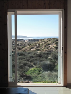 A room with a sliding glass door leading to a balcony with a view of a hilly landscape and the ocean.