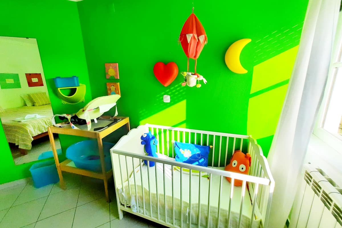 A room with a crib, a bed, and a desk with a chair. There are wall decorations including a heart, a crescent moon, and a hanging ornament.