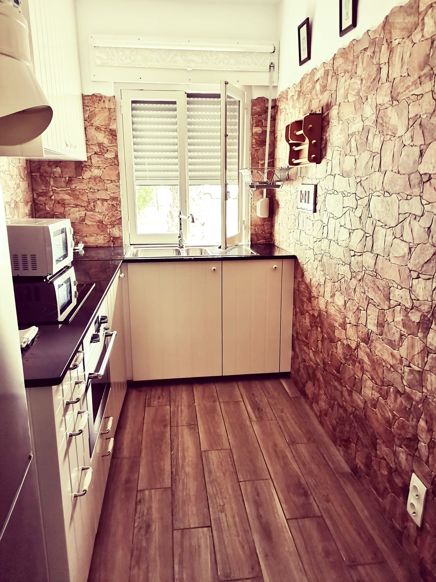 A compact kitchen with stone walls, a built-in microwave, and a window with blinds.