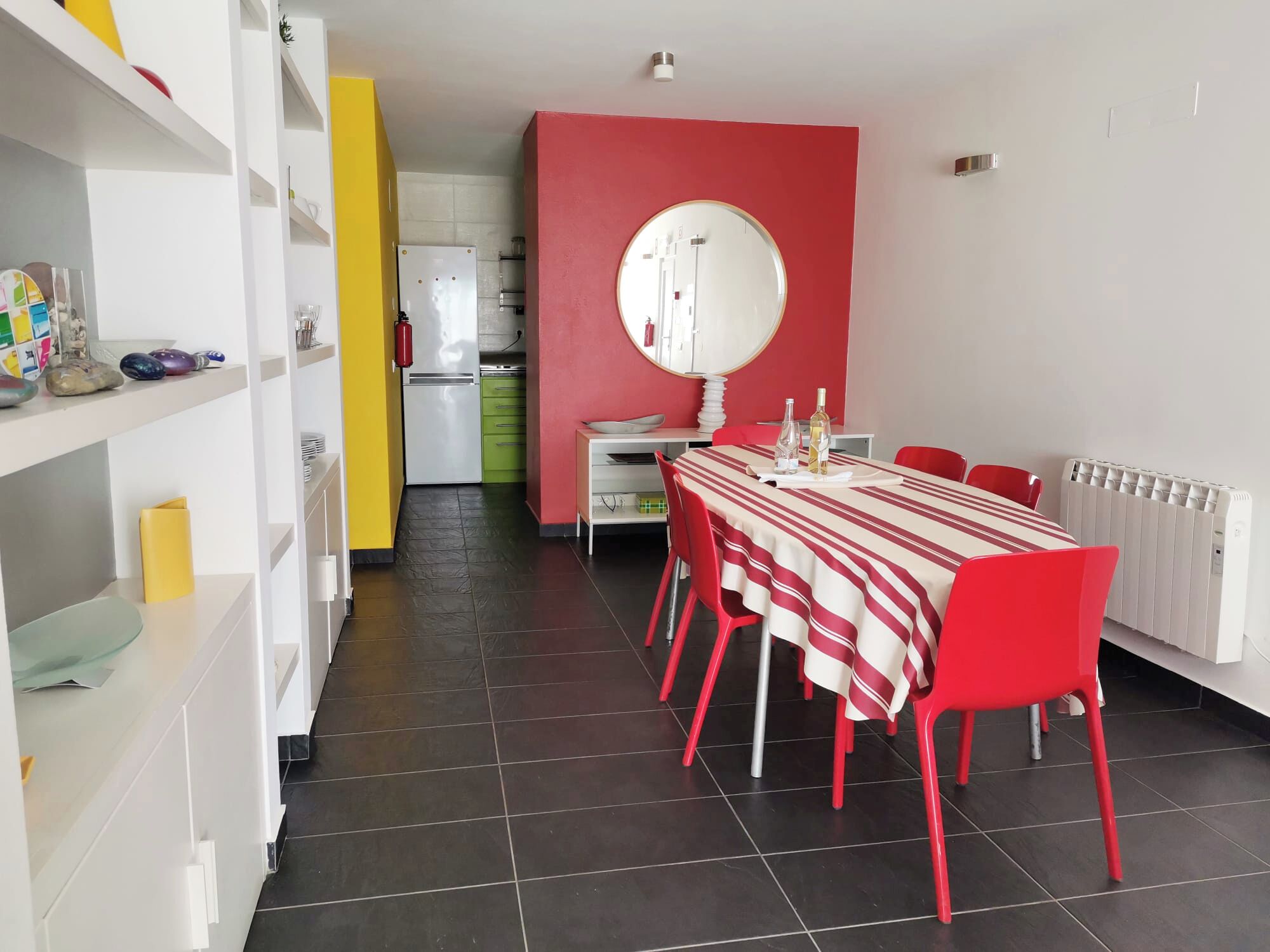Uma área de jantar moderna com toalha de mesa listrada em vermelho e branco, cadeiras vermelhas e um espelho redondo grande na parede.