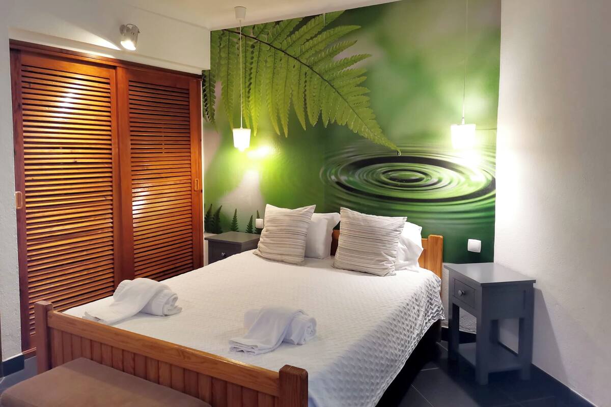 A neatly made bed with white linens and pillows, a wooden headboard, a nightstand with a lamp, and a wall with a green leaf mural.