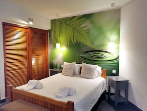 A neatly made bed with white linens and pillows, a wooden headboard, a nightstand with a lamp, and a wall with a green leaf mural.