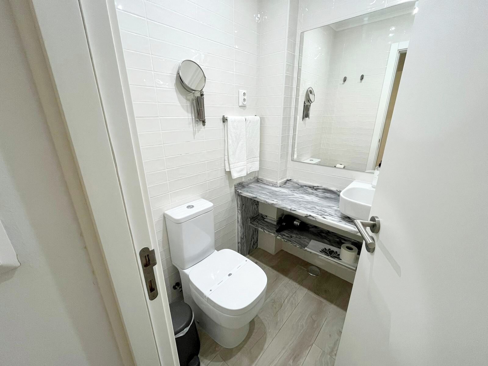 A modern bathroom with white tiles, a marble countertop, a toilet, and a mirror.