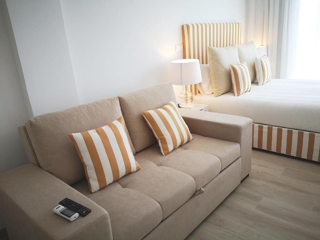 Habitación de hotel con sofá color beige, almohadas a rayas, una mesita de noche con lámpara y cama con ropa de cama a rayas.