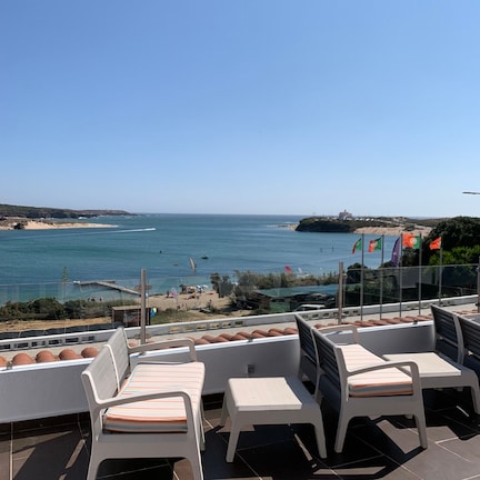 A terrace with white plastic chairs and a table, overlooking a coastal area with flags and a beach.