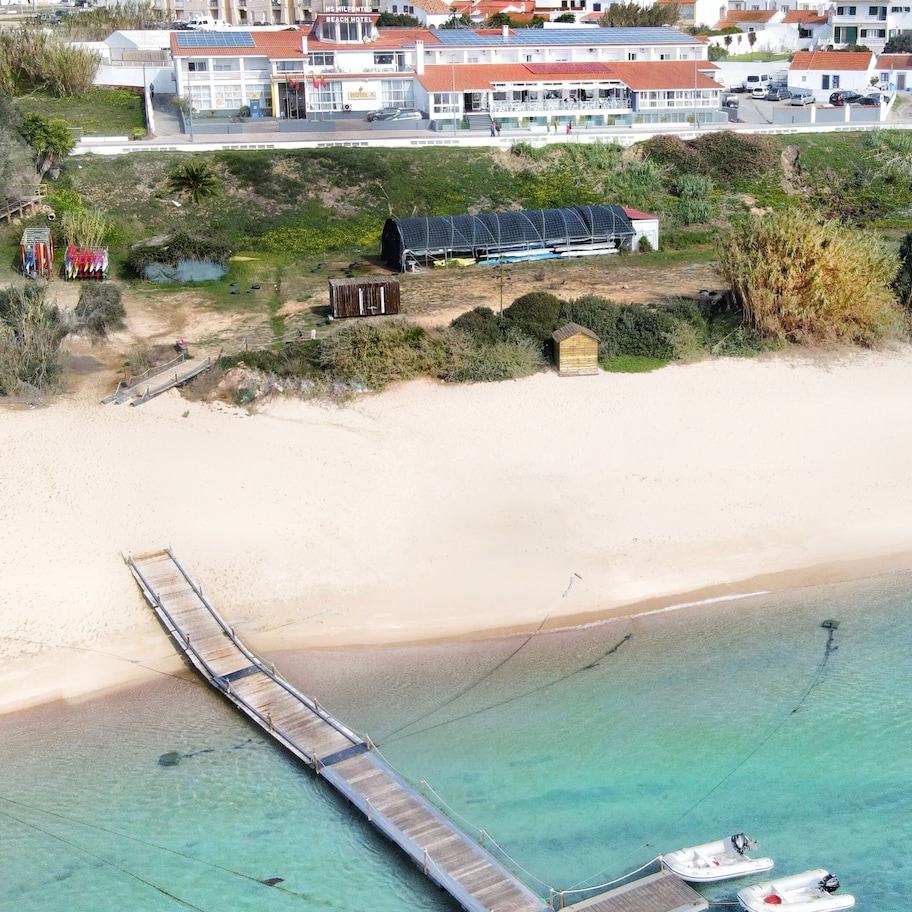 An aerial view of a coastal area with a pier extending into turquoise waters, a small wooden hut, and a building with solar panels.