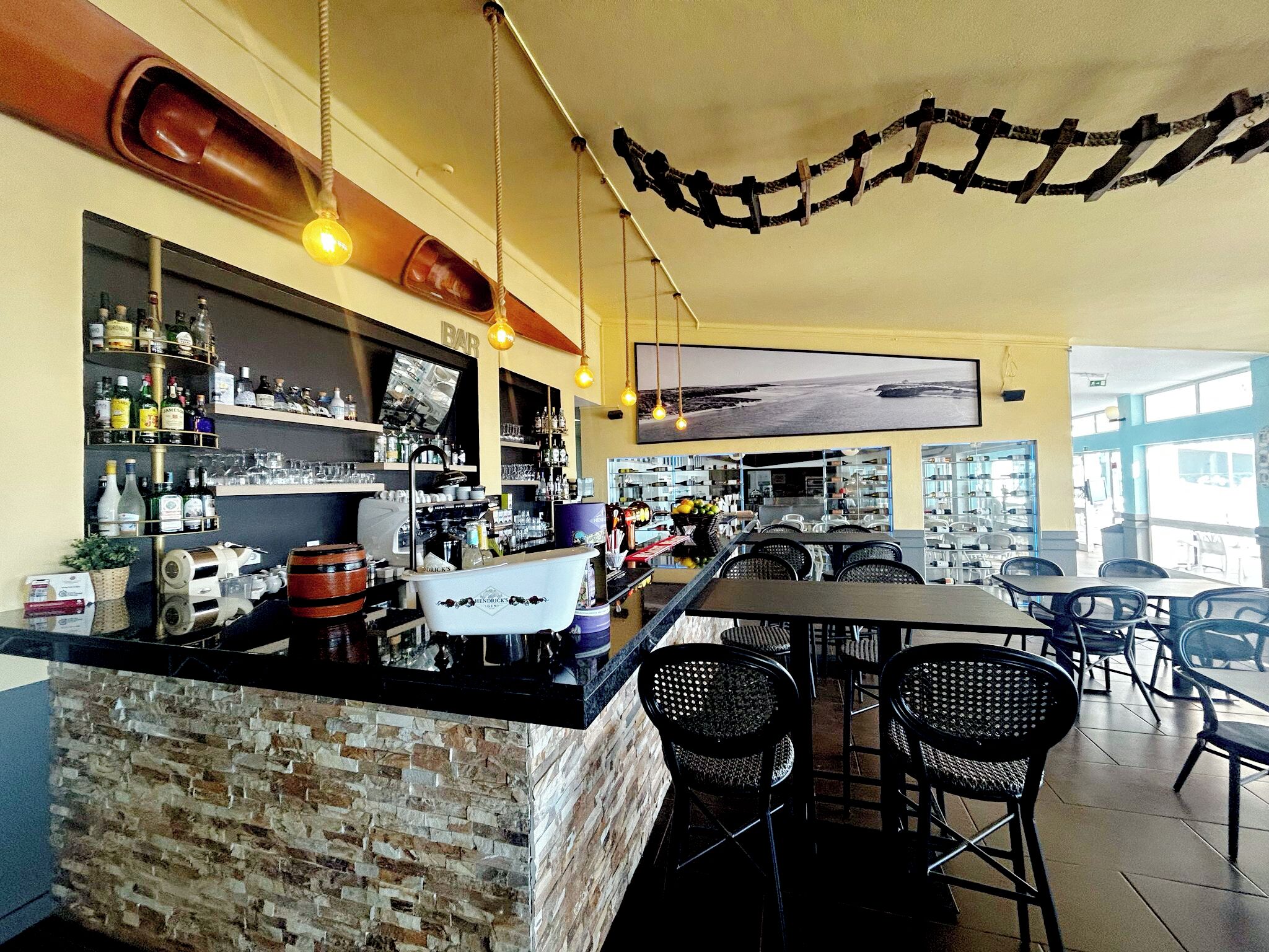 A bar area with a brick counter, shelves of bottles, and hanging lights. There are tables and chairs for seating.