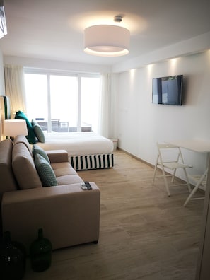 A modern hotel room with a large bed, a sofa, a desk, and a TV on the wall.