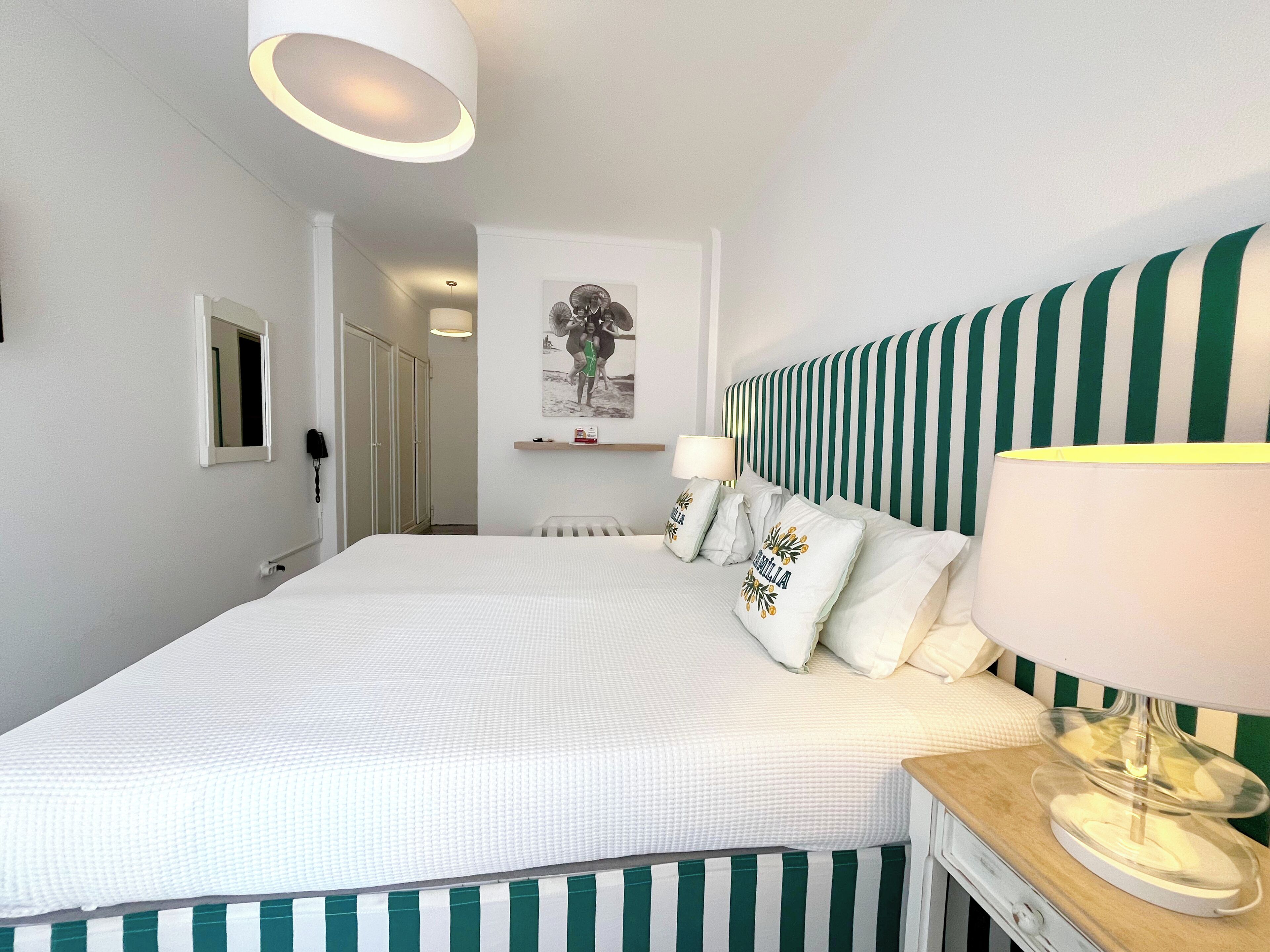 A hotel room with a large bed, a striped headboard, a bedside table with a lamp, and a wall-mounted shelf with decorative pillows.